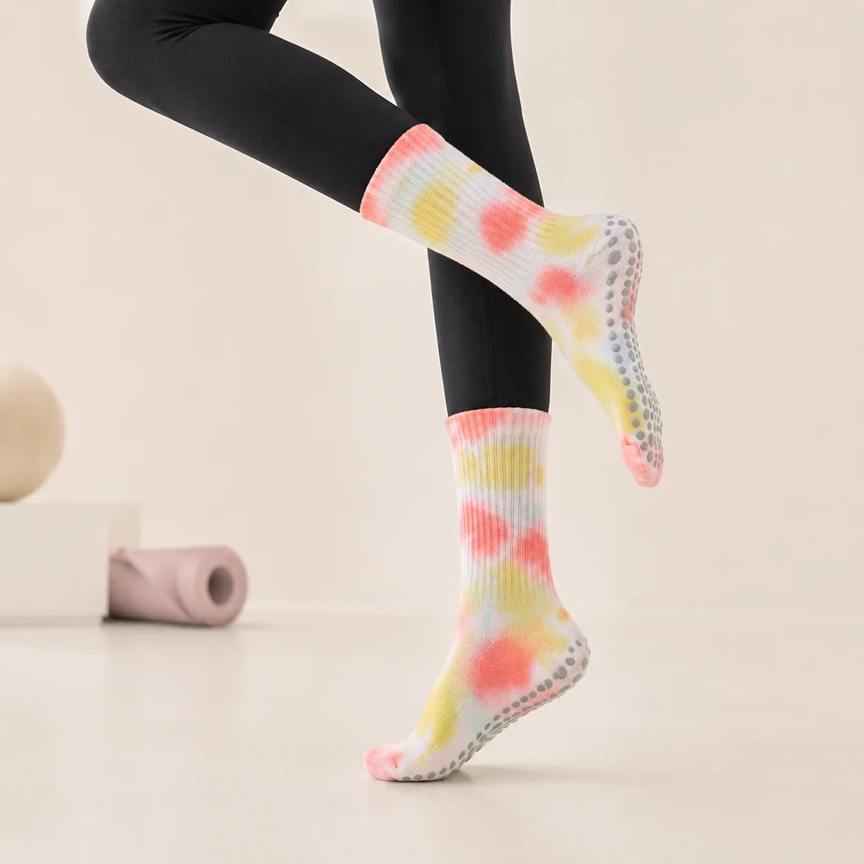 A person wearing black leggings and colorful tie-dye grip socks with gray non-slip pads, shown against a clean light beige studio background.