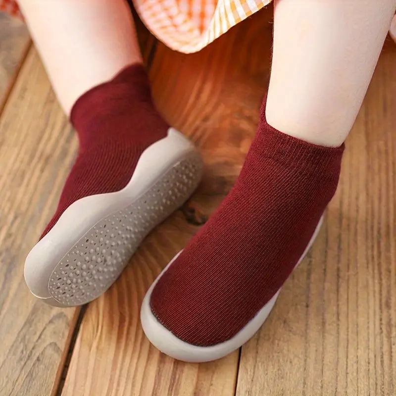 Child wearing burgundy Mini Walker sock shoes with flexible non-slip soles on a wooden floor