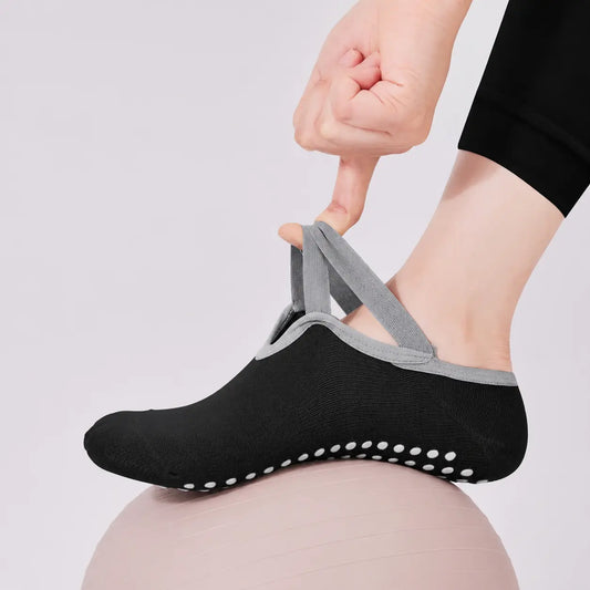 Black Pilates grip sock with grey crossover straps being stretched by hand on an exercise ball