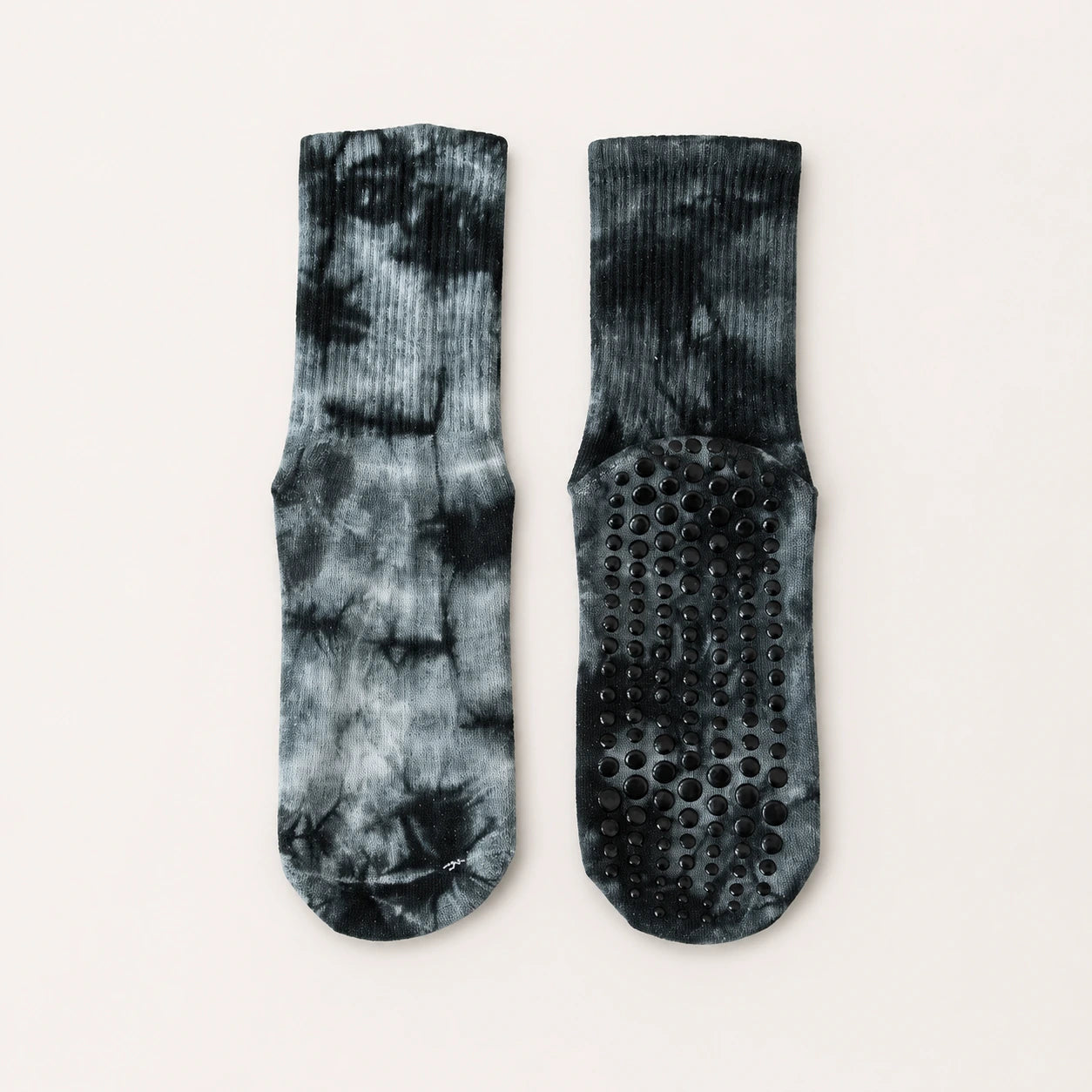 A pair of crew socks with a black and gray tie-dye pattern, shown from both the front and back. The back of the socks features a black grip sole with raised circular dots for traction, set against a plain off-white background.