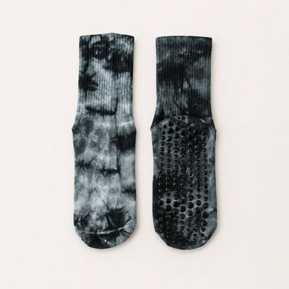 A pair of crew socks with a black and gray tie-dye pattern, shown from both the front and back. The back of the socks features a black grip sole with raised circular dots for traction, set against a plain off-white background.