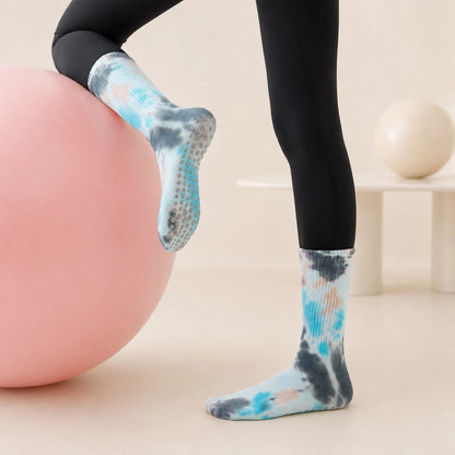 A person wearing black leggings and blue, gray, and white tie-dye grip socks stands on a pink exercise ball in a clean studio setting.