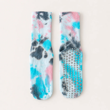 A pair of ribbed grip socks with a tie-dye pattern in turquoise, pink, and charcoal gray on a white base, displayed flat against a light beige background.