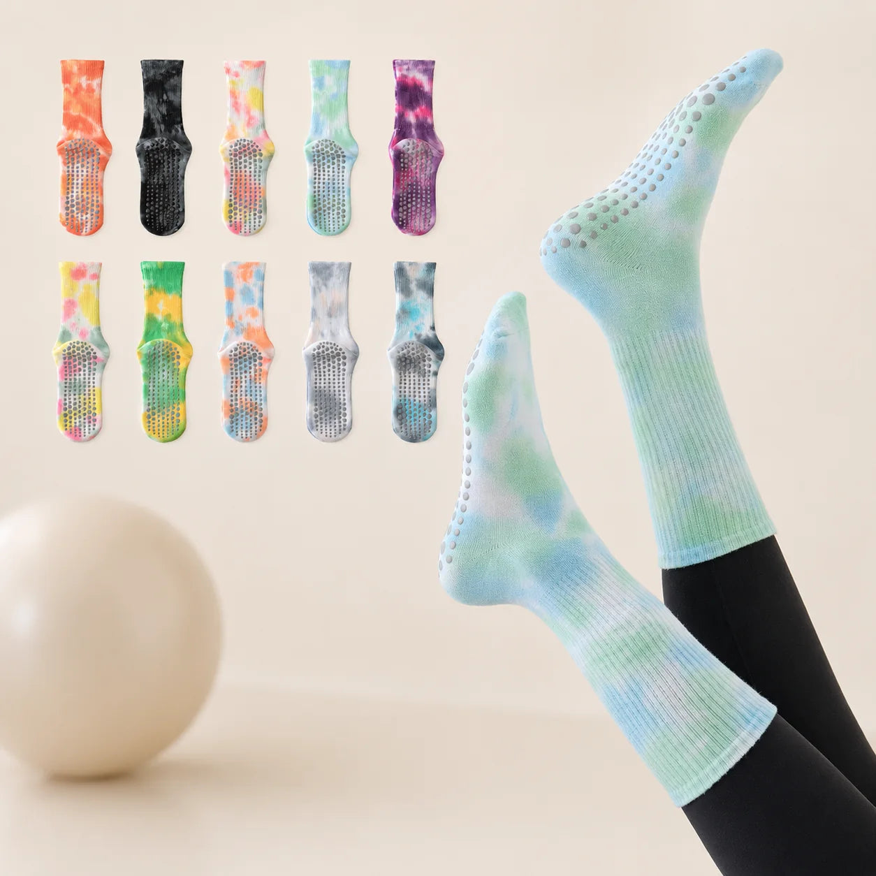 Tie-dye grip socks worn on feet with black leggings, shown against a warm beige background with a small decorative sphere. Eight additional tie-dye sock options are displayed in a grid at the upper left.