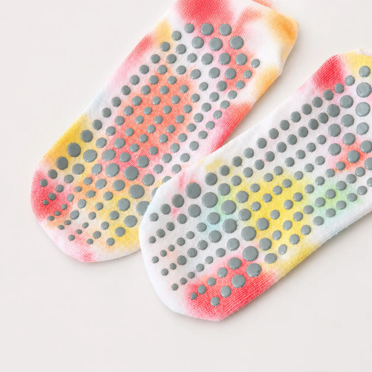 A pair of white grip socks with a colorful tie-dye pattern in pink, orange, yellow, and green, featuring gray silicone non-slip dots on the sole, shown on a light beige background.