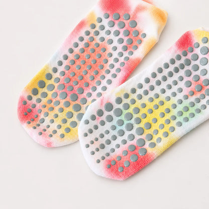 A pair of white grip socks with a colorful tie-dye pattern in pink, orange, yellow, and green, featuring gray silicone non-slip dots on the sole, shown on a light beige background.