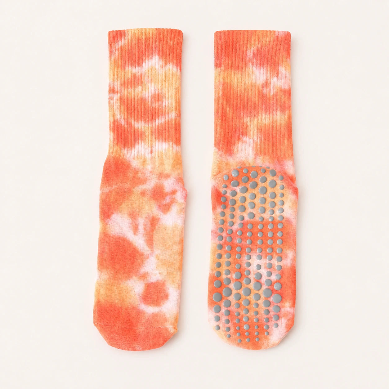 A pair of tie-dye grip socks in orange, peach, and white, displayed flat against a light cream background. One sock is shown from the side, highlighting the ribbed cuff, while the other is shown from the sole, displaying the textured non-slip grip dots.