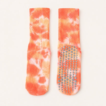 A pair of tie-dye grip socks in orange, peach, and white, displayed flat against a light cream background. One sock is shown from the side, highlighting the ribbed cuff, while the other is shown from the sole, displaying the textured non-slip grip dots.