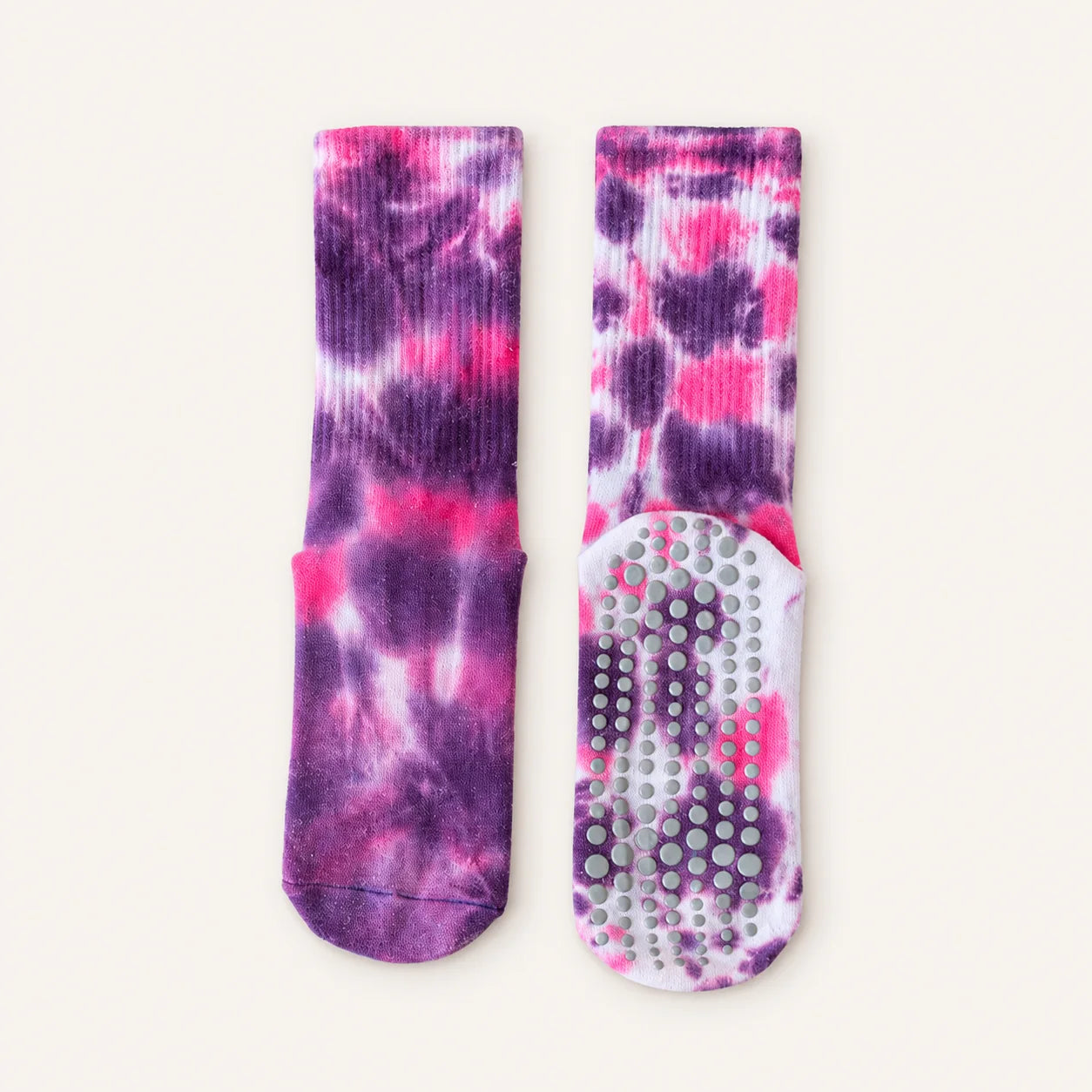 A pair of purple and pink tie-dye socks, one showing the ribbed knit upper and the other showing the textured non-slip grip sole, laid flat on a light cream background.